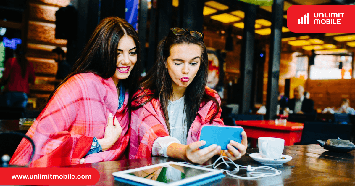 Two friends in a café looking at a smartphone together, with a tablet and a coffee cup on the table