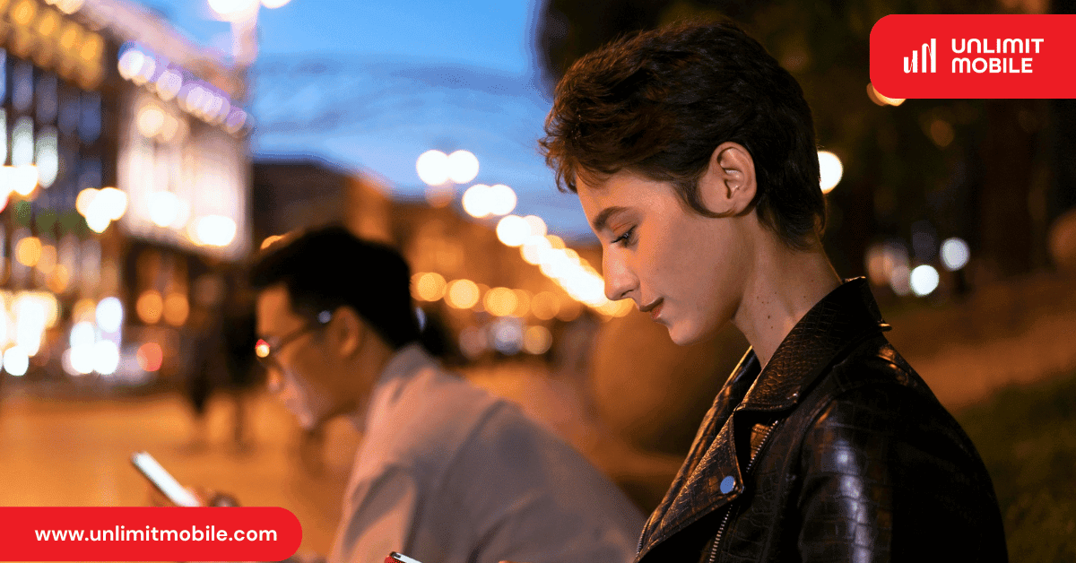 Two people checking their phones on a city street lit up at night